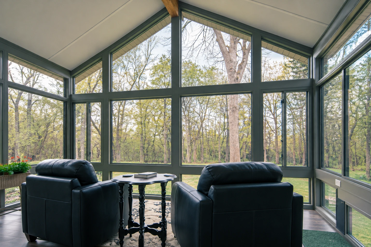Four-season sunroom with dark-framed floor-to-ceiling windows, two black armchairs, and a wooded view.