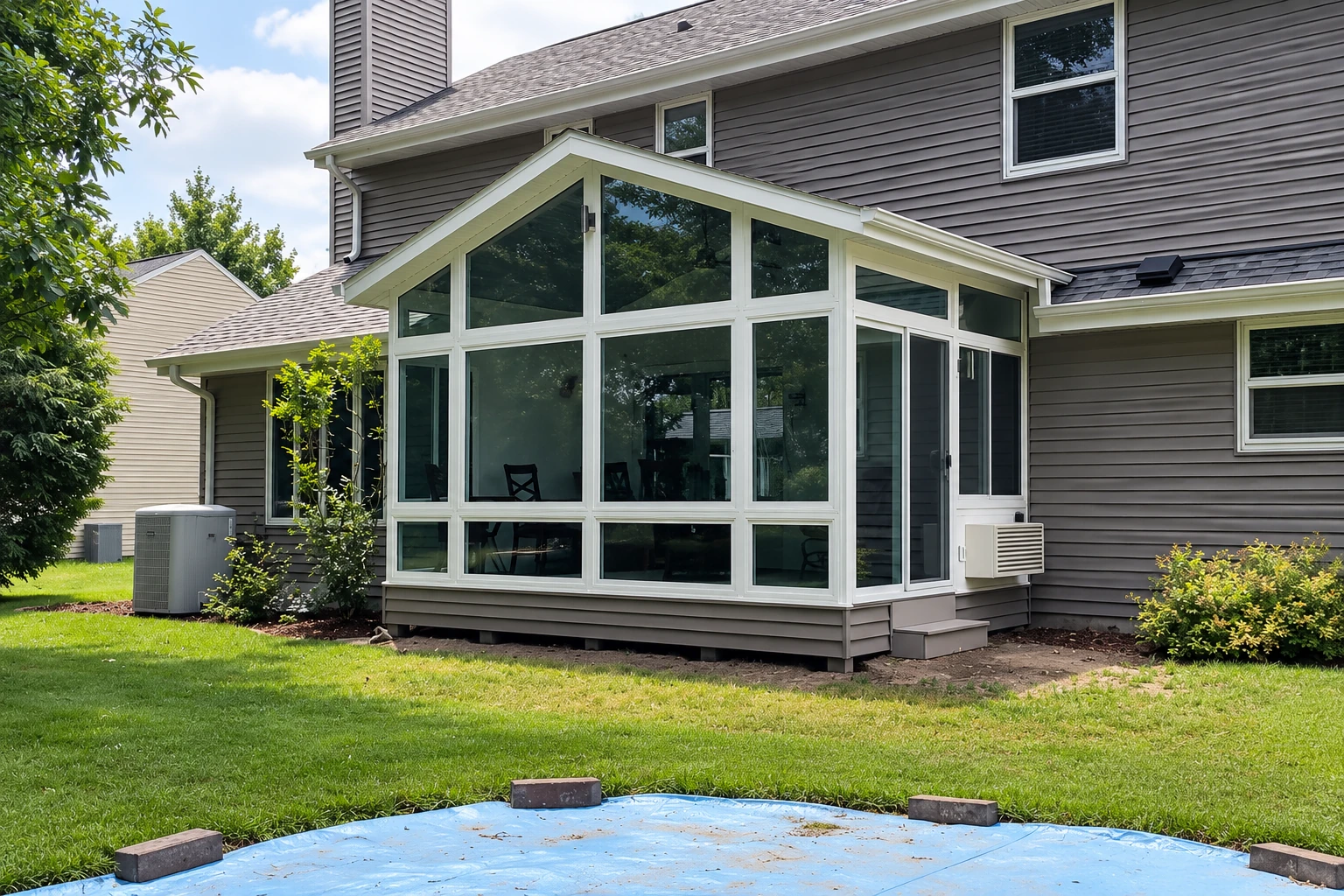 White-framed, gabled glass sunroom attached to a gray house overlooks a lawn, with unfinished paving visible in front.