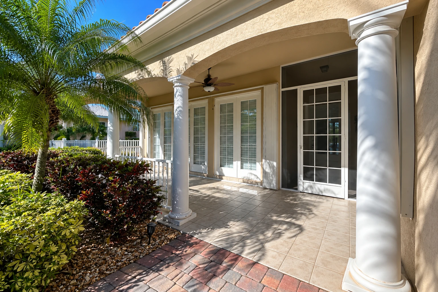 Covered patio with white columns, tiled floor, ceiling fan, and glass doors beneath a stucco arch, surrounded by tropical plants.