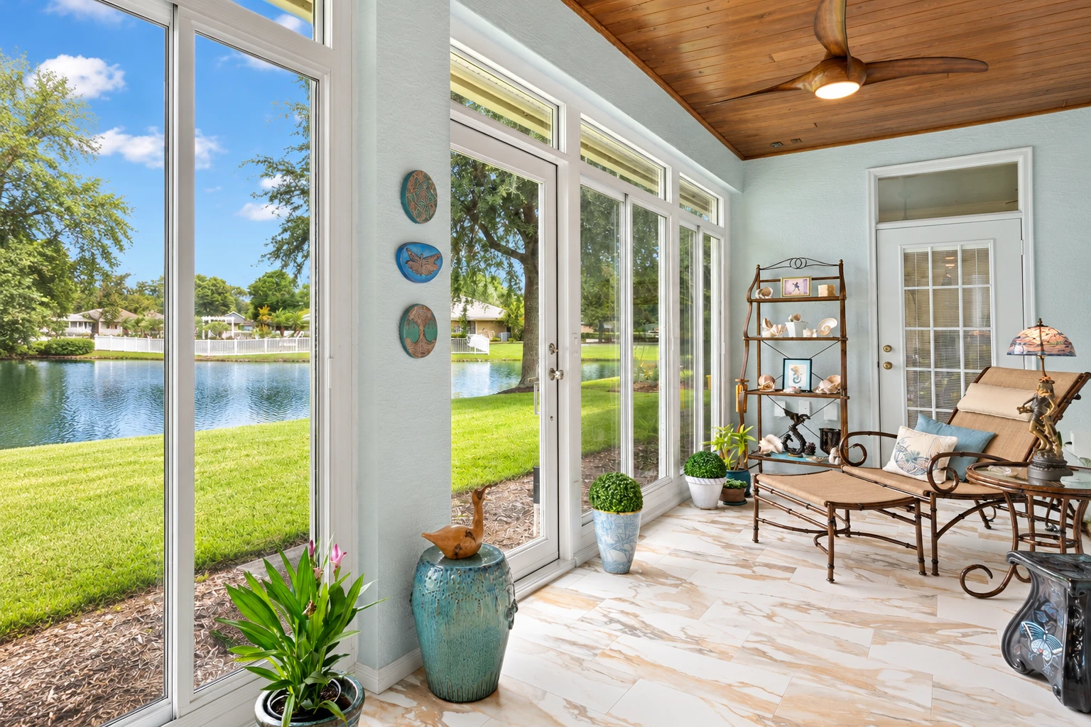 Sunroom addition with tall white-framed windows overlooking a lake, furnished with wicker seating beneath a wood-paneled ceiling.
