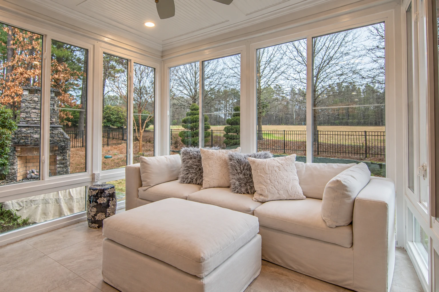 Bright sunroom interior with tall windows, a cream sofa and ottoman, and a view of trees and a fenced yard.