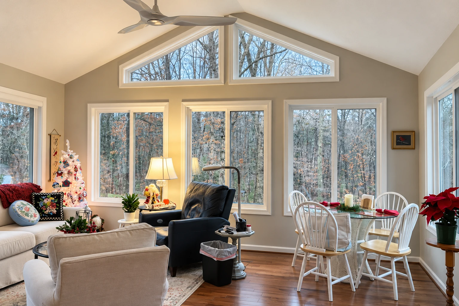 Sunroom with high triangular clerestory windows, a ceiling fan, seating and a dining table beside winter trees.