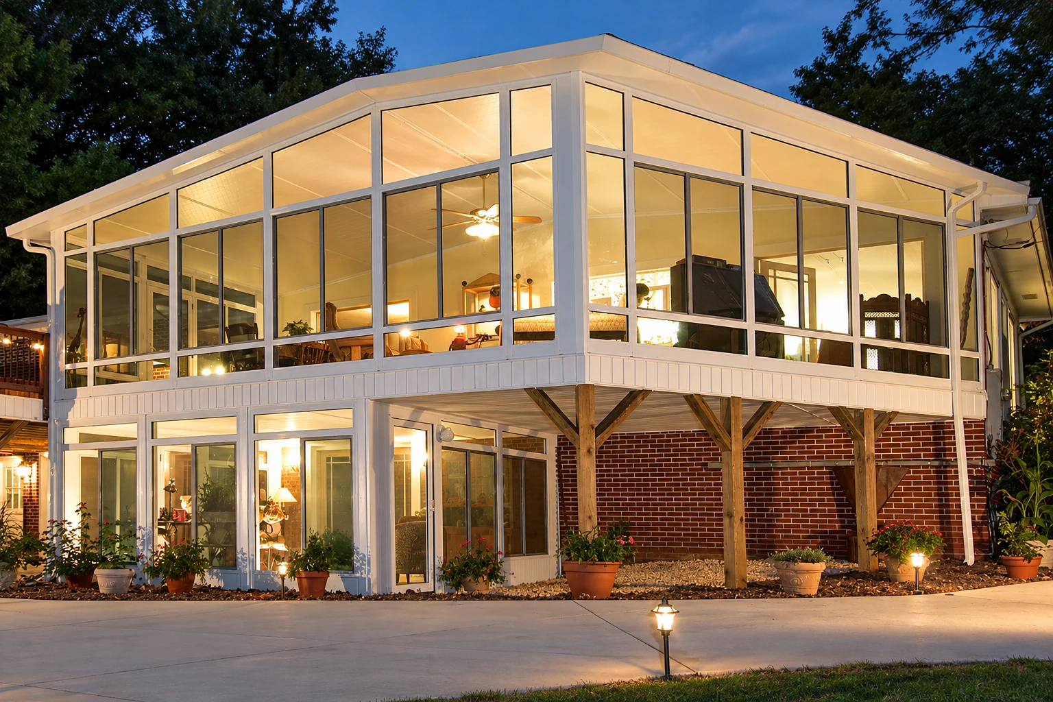 Raised three-season sunroom with broad white-framed windows glowing at dusk above a brick-and-wood-supported lower level.