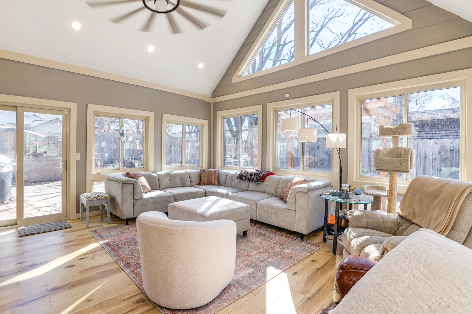 Bright sunroom living area with vaulted ceiling, tall windows, sectional sofa and wood floors.
